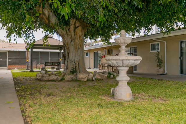 Outdoor courtyard area at Rose Villa Healthcare Center featuring a large tree surrounded by rocks, a stone fountain, green grass, and a building with multiple doors and windows in the background.