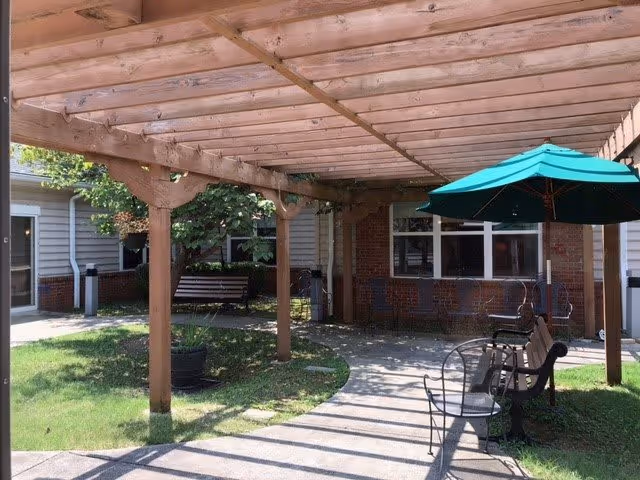 Outdoor seating area at The Rutherford Memory Care featuring a wooden pergola, metal benches, a green patio umbrella, and a paved walkway surrounded by grass and plants.