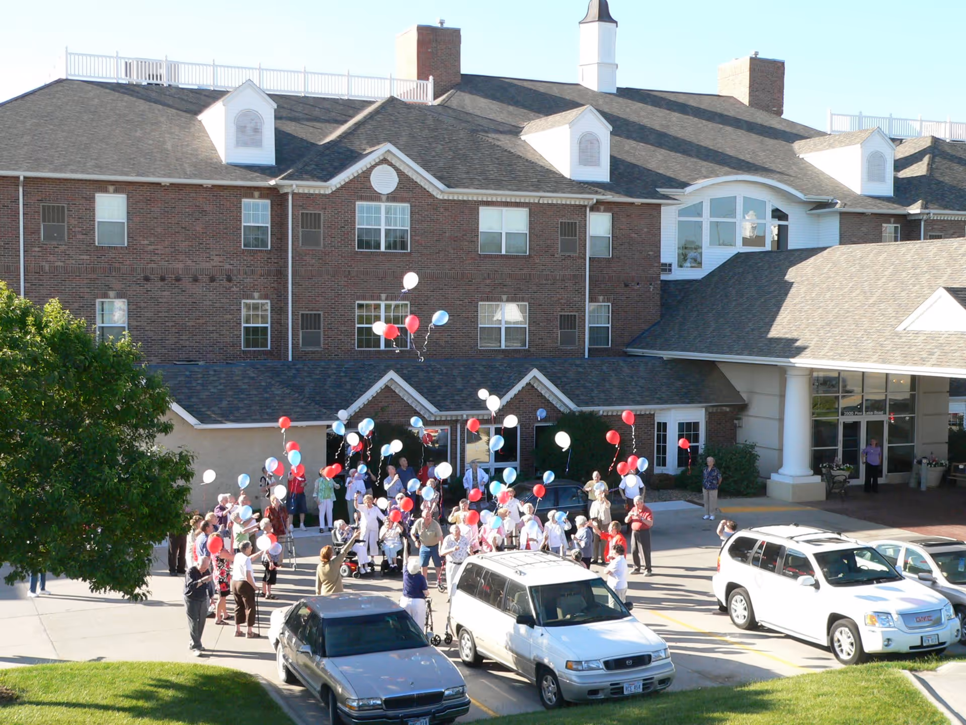 A group of people gathered outside a large brick retirement community building, releasing red, white, and blue balloons into the air on a sunny day. Several cars are parked in front of the building, and a tree is visible on the left side.