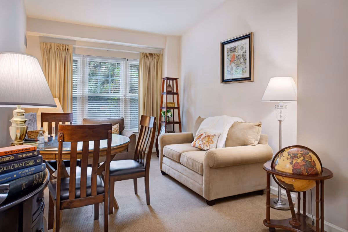 Cozy living room and dining area featuring a beige loveseat, round wooden dining table with chairs, floor and table lamps, a globe on a stand, and a window with blinds and curtains.