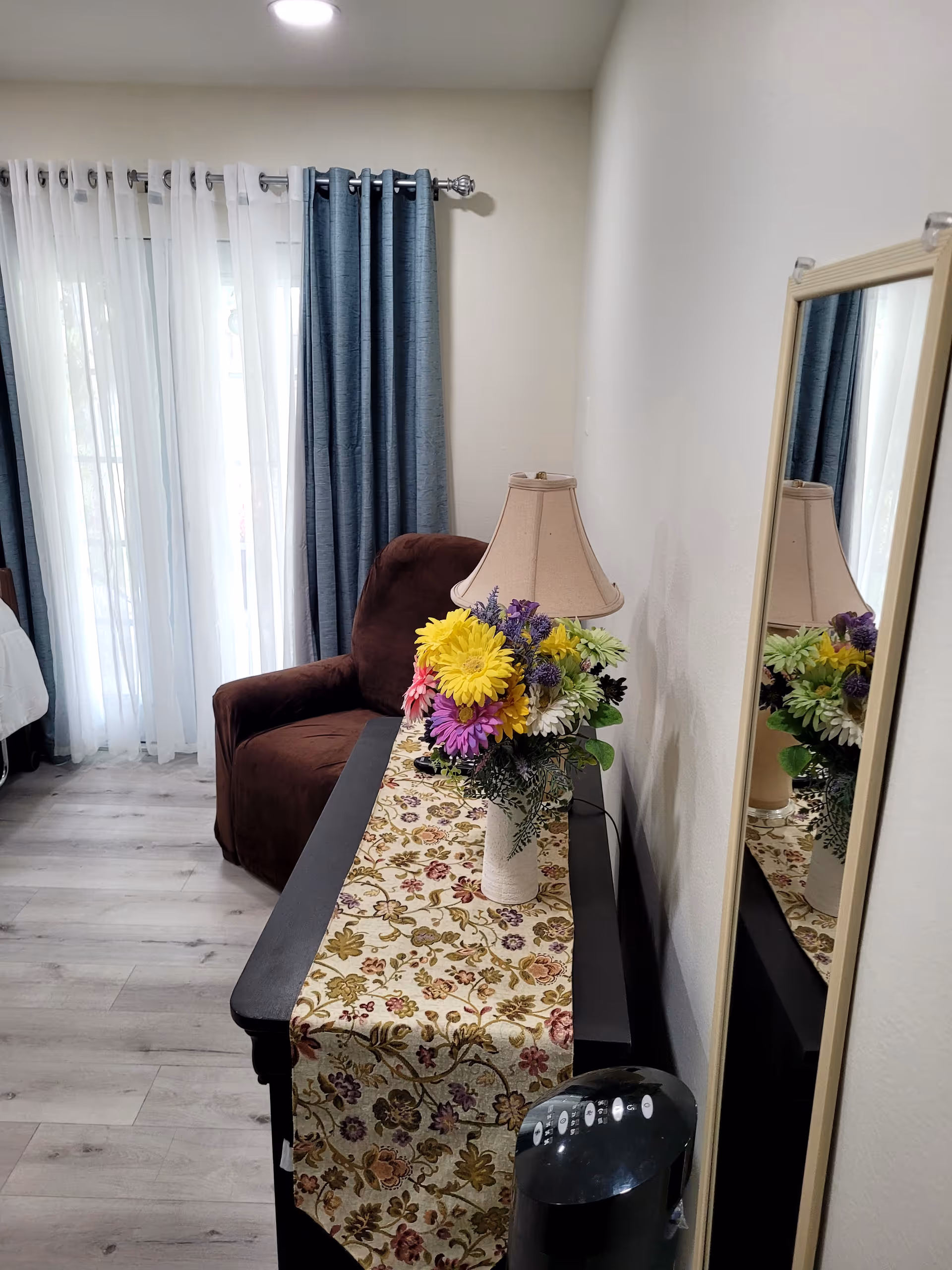 A cozy interior room with a brown armchair next to a window covered by sheer white and blue curtains. A black table with a floral table runner holds a vase of colorful flowers and a beige table lamp. A tall mirror is mounted on the wall reflecting the flowers and lamp. The floor is light wood.