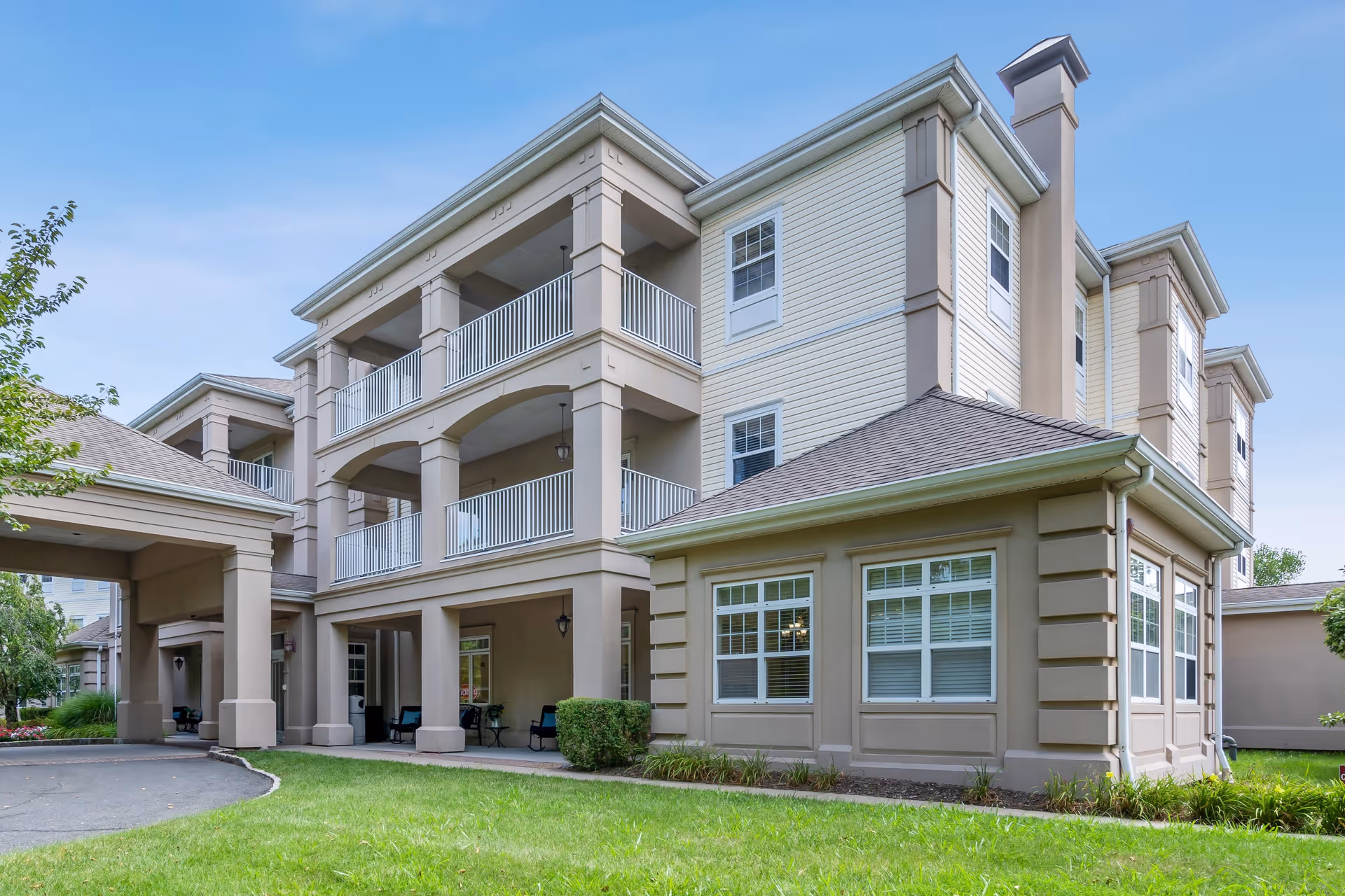 Exterior view of a multi-story assisted living facility building with beige and light tan siding, multiple balconies with white railings, and a covered entrance. The building is surrounded by green grass and some trees under a clear blue sky.
