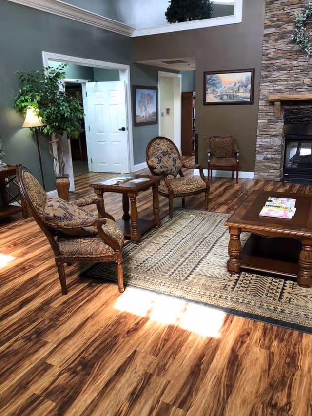 A cozy senior living facility common area with wooden flooring, a patterned area rug, and classic upholstered chairs around wooden tables. There is a stone fireplace on the right wall with a framed painting above it, and a tall green plant with a floor lamp on the left side near an open white door. The walls are painted in muted green and brown tones.