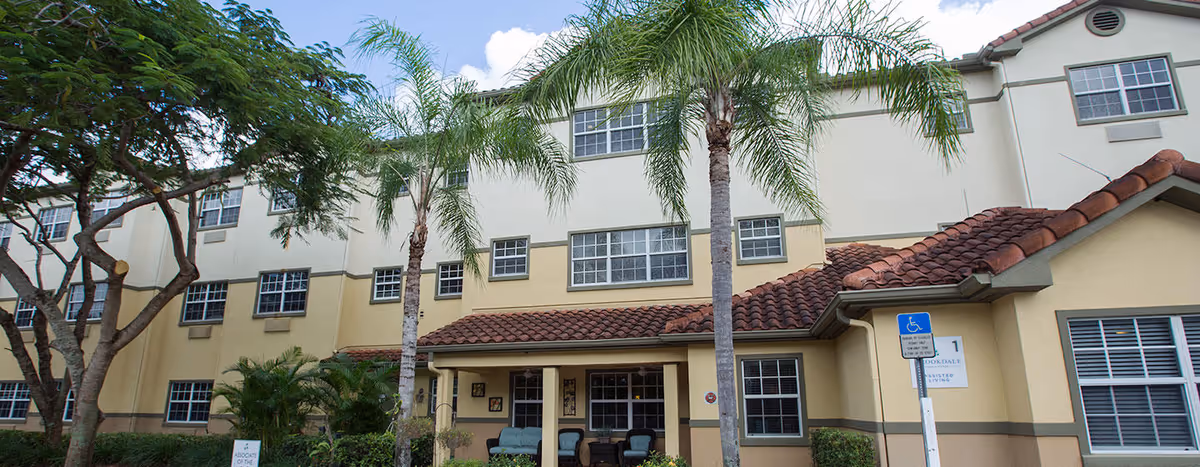 Exterior view of a multi-story senior living facility with beige and yellow walls, red-tiled roof, multiple windows, palm trees, and a handicap parking sign near the entrance.