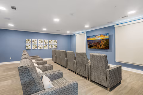 A small theater-style common room with two rows of upholstered recliners facing a wall-mounted flat-screen TV and blank projector screens on blue walls.