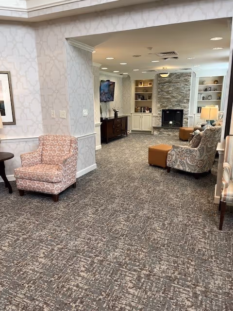Carpeted lounge area with patterned armchairs, an ottoman, built-in shelves and a stone fireplace in a senior living facility interior.
