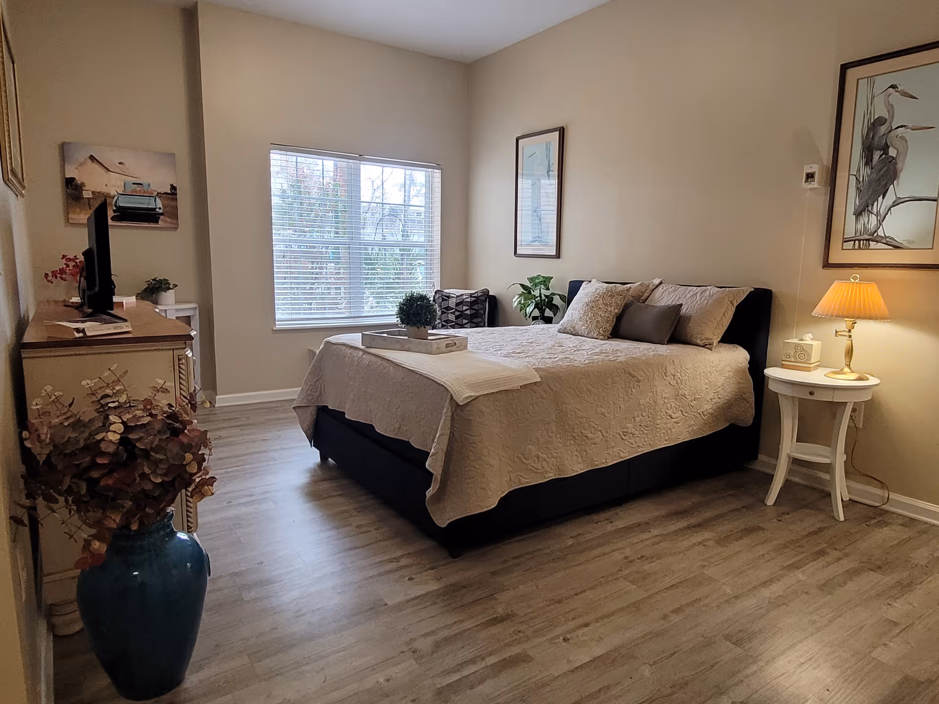 A cozy bedroom with a large bed covered in a beige quilt and multiple pillows. There is a window with blinds letting in natural light, a wooden dresser with a TV on top, a small white side table with a lamp, and framed artwork on the walls. The floor is light wood, and there is a blue vase with dried flowers in the corner.