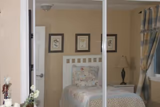 A cozy bedroom with a white bed frame, a bed covered with a patchwork quilt and pillows, a nightstand with a lamp, three framed pictures on the wall, and a window with plaid curtains.