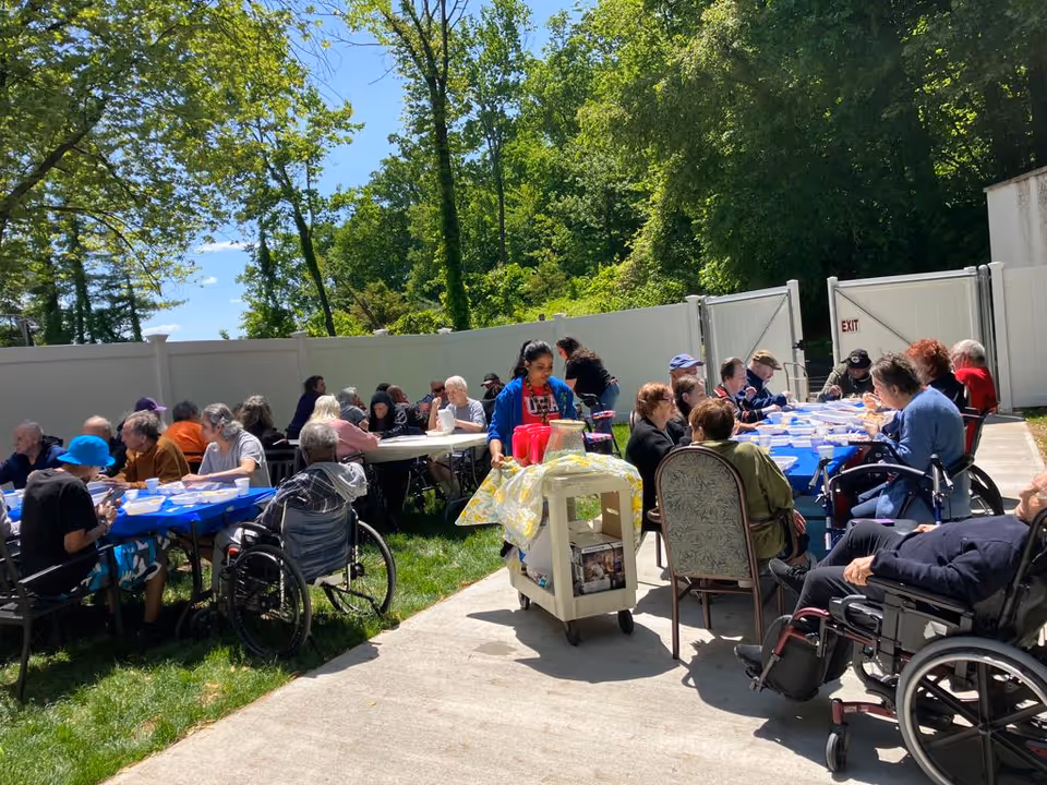 A group of elderly people sitting outdoors at tables covered with blue tablecloths, enjoying a meal together on a sunny day. Some individuals are in wheelchairs, and a caregiver is serving drinks from a cart. The area is surrounded by a white fence and trees with green foliage under a clear blue sky.