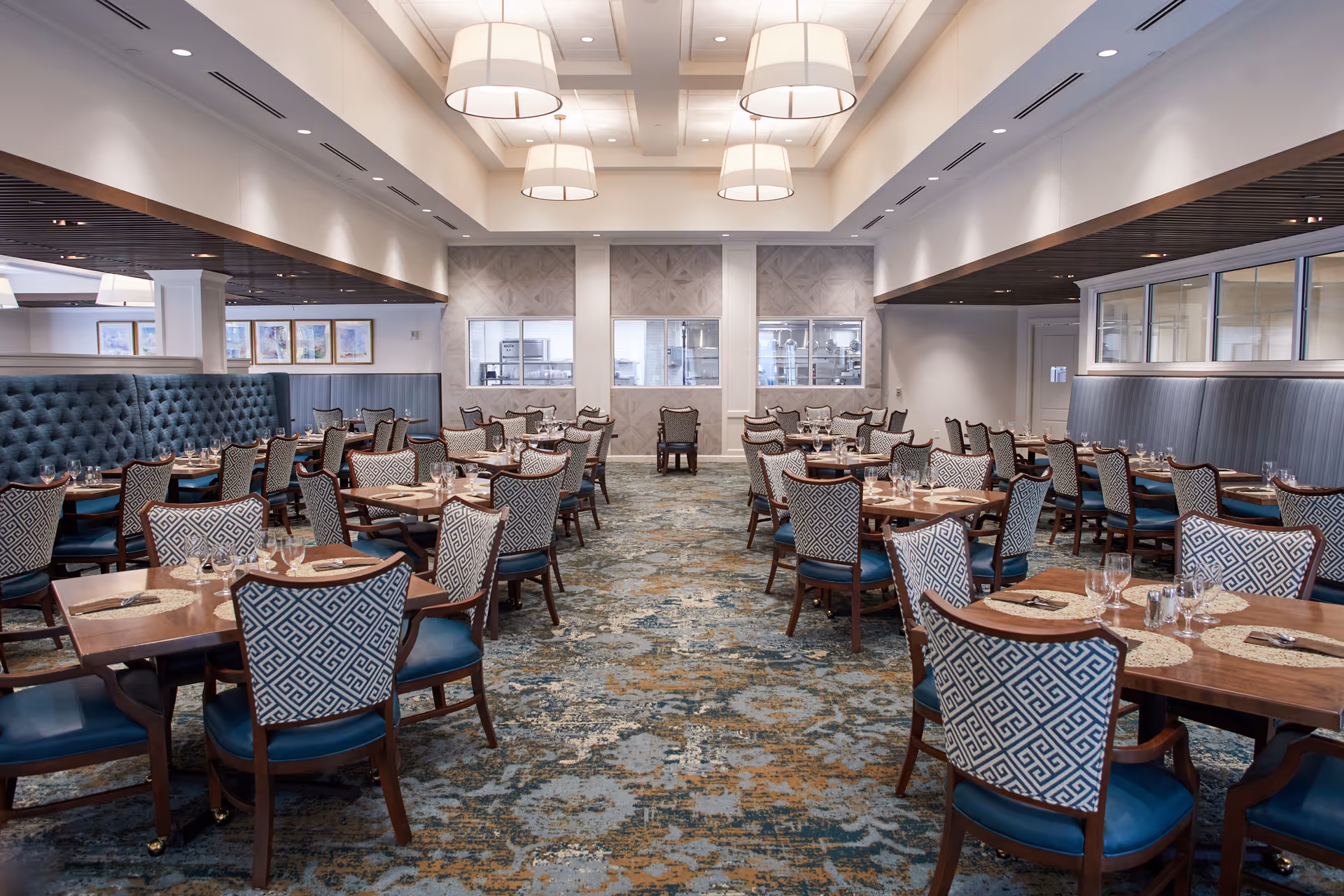 A spacious and elegant dining room with multiple wooden tables and patterned upholstered chairs arranged neatly. The room features a patterned carpet, large pendant lights hanging from the ceiling, and a tufted blue banquette along one side. The far wall has windows looking into a kitchen area.