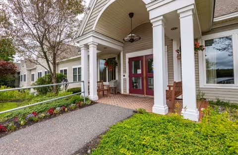 Covered building entrance with double red doors, white columns, seating, a ramp, and landscaped greenery.