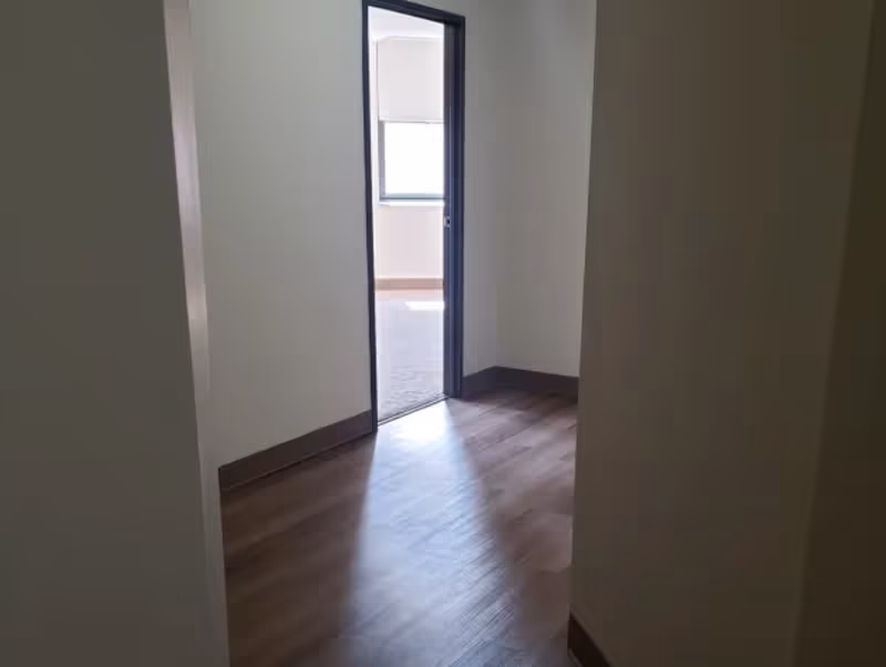 View of a hallway with wooden flooring leading to a room with a window letting in natural light. The walls are plain and light-colored with dark baseboards.