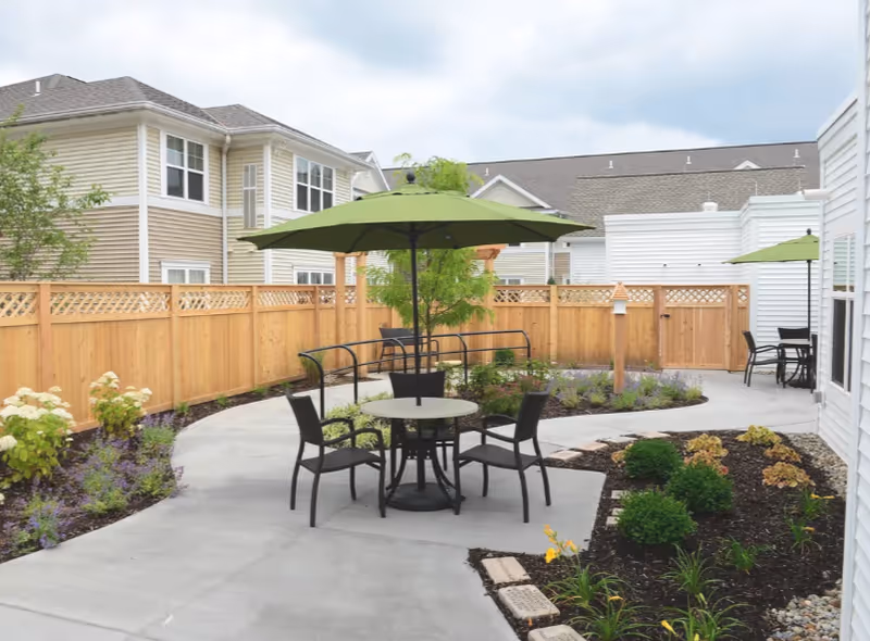 Outdoor patio area with a round table and four chairs under a green umbrella, surrounded by landscaped garden beds and a wooden fence, with residential buildings in the background.