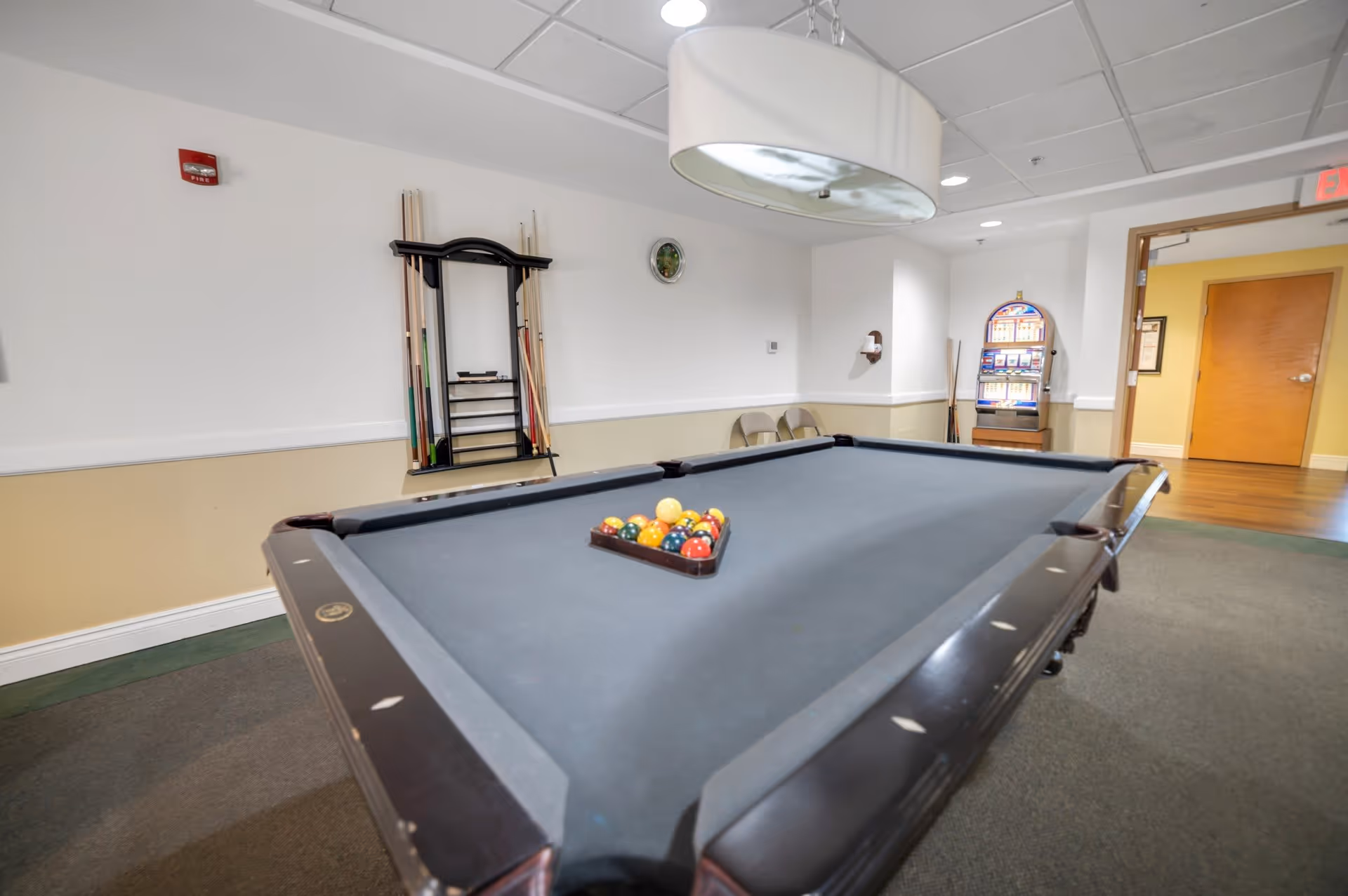 Interior room with a pool table set up for a game, pool cues on a rack against the wall, two folding chairs, a wall clock, a hanging light fixture above the table, and an open doorway leading to a hallway with wooden doors.