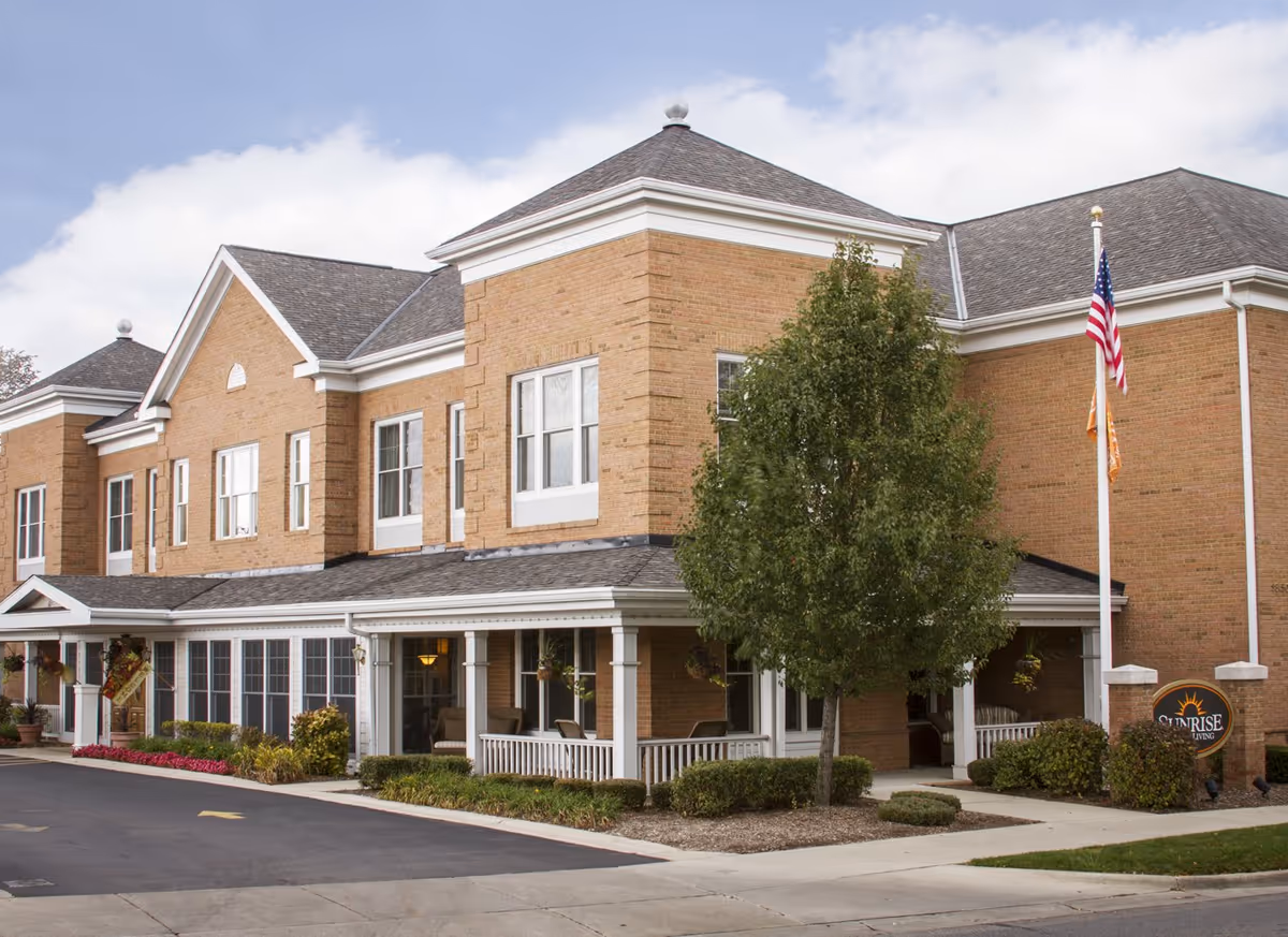 Exterior view of a two-story brick building with white trim, a covered porch with seating, landscaping including bushes and a tree, and two flagpoles displaying the American flag and another flag. A sign near the entrance reads 'Sunrise Living'.