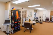A well-lit room in a senior living facility with exercise equipment including stationary bikes and a rack of colorful dumbbells. There is a table with chairs in the center, a laptop on a small table, and a cabinet with glass doors. The room has beige walls and carpeted floors.