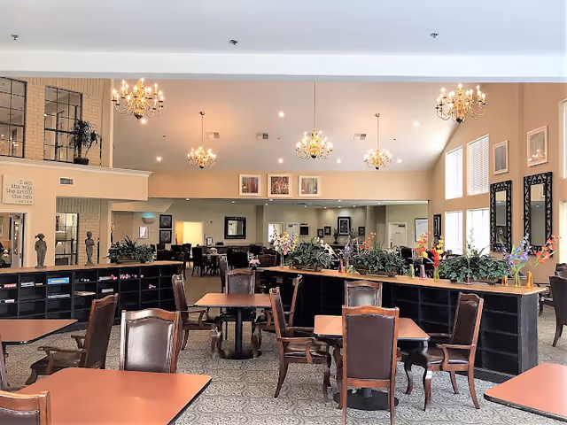 Open dining room in a senior living facility with wooden tables and chairs, chandeliers, plants, and large windows.