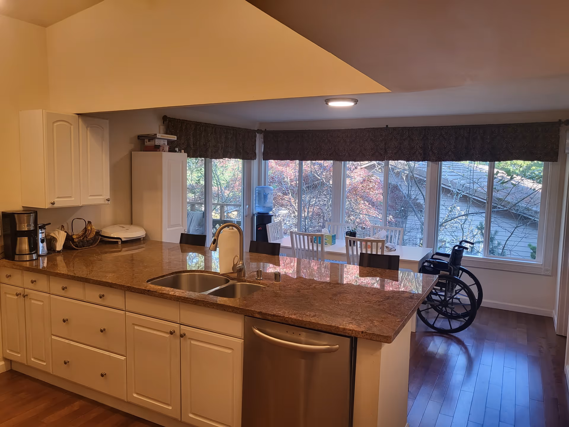 Interior view of a kitchen area with a large countertop island featuring a double sink and dishwasher. In the background, there is a dining area with a white table, several chairs, a water dispenser, and a wheelchair near large windows with a view of trees outside.