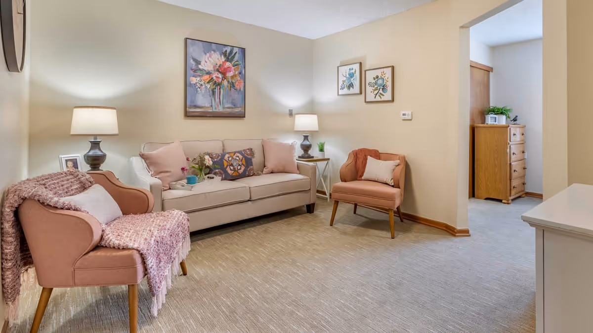A cozy living room with a beige sofa adorned with pink and patterned cushions, two pink armchairs with a pink throw blanket, two table lamps on side tables, and floral artwork on the walls. The room has light-colored carpet and beige walls, with an open doorway leading to another room with wooden furniture.