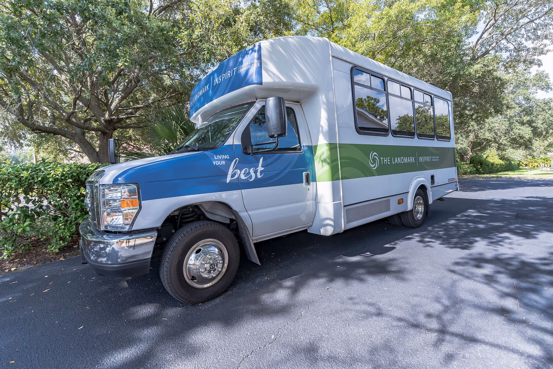 A white and blue shuttle bus with green stripe parked on a paved driveway surrounded by trees and bushes. The bus has text on the side that reads 'THE LANDMARK' and 'LIVING YOUR best'.