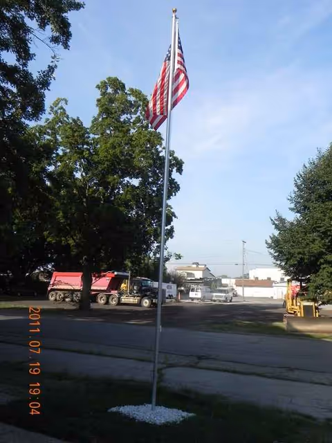 American flag on a tall pole in a parking lot with trees and trucks.