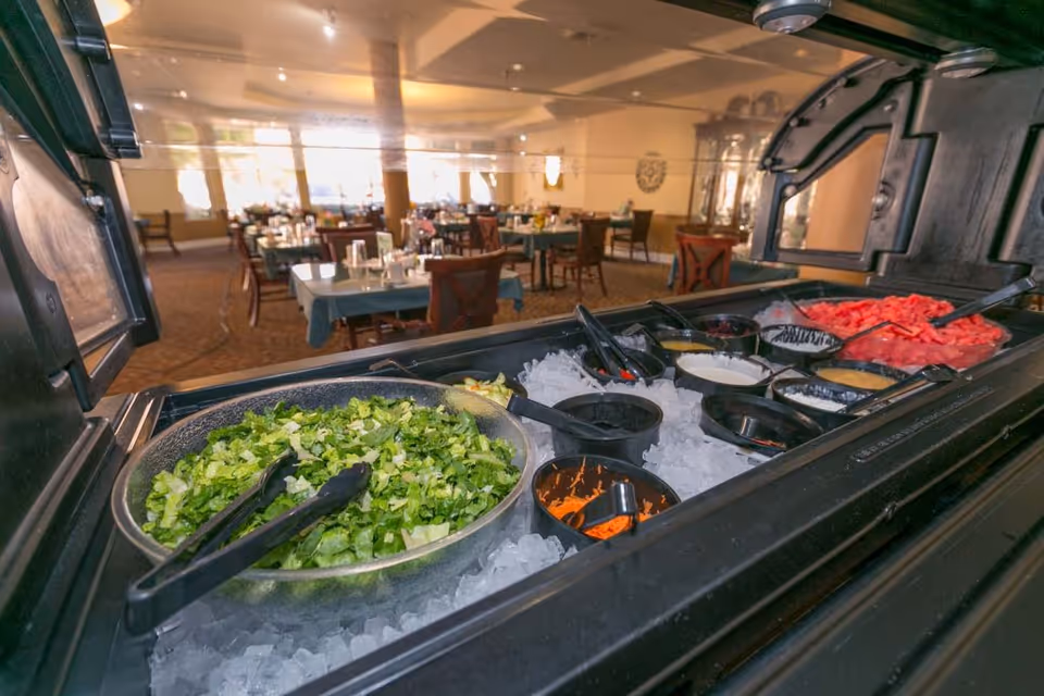Salad bar with bowls of chopped lettuce, dressings and toppings on ice, with a dining room and set tables in the background.