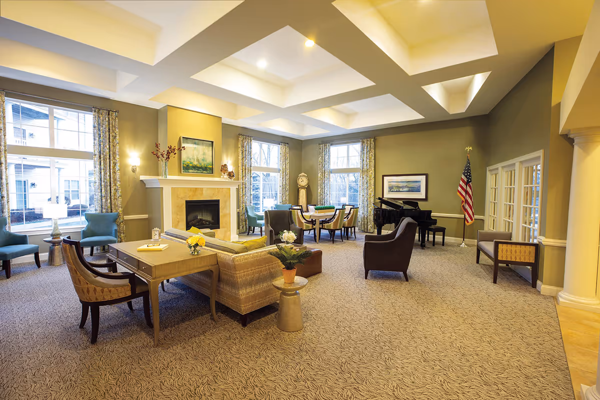 A spacious and well-lit living room area with a beige carpet and light yellow walls. The room features a fireplace with a painting above it, several chairs and sofas arranged around a wooden table, a grand piano in the corner, an American flag, and large windows with patterned curtains allowing natural light to enter.