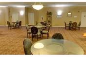 A spacious dining room with multiple round tables and chairs arranged throughout the room. The carpet has a patterned design, and the walls are painted a light color with several wall-mounted lights providing illumination. There is a decorative shelving unit against the far wall and a door visible in the background.
