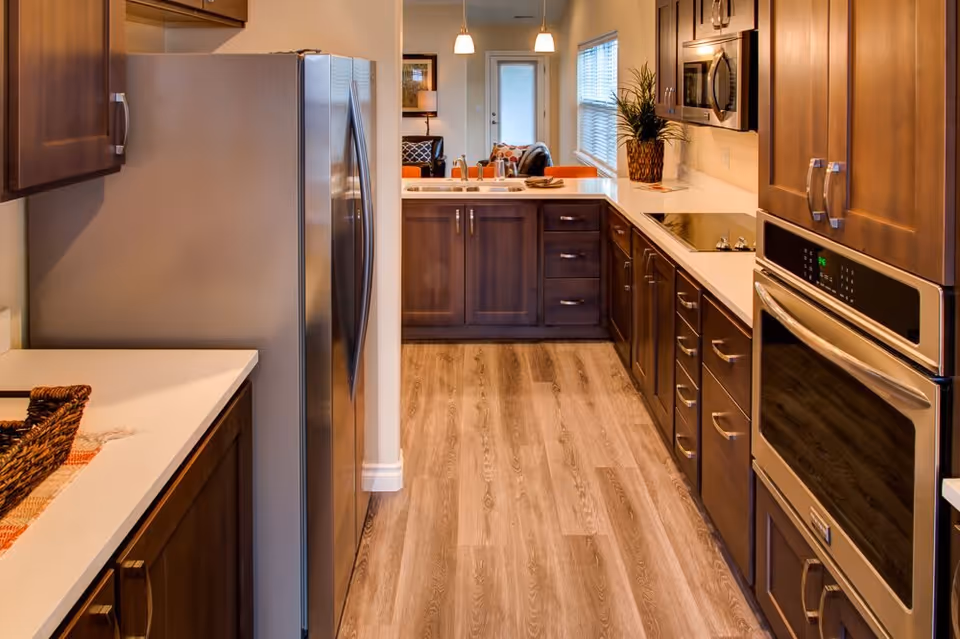 A modern kitchen with wooden cabinets, stainless steel refrigerator, built-in oven, microwave, and electric cooktop. The kitchen has light-colored countertops and wood flooring, with a view into a living area with a sofa and a door with a window.