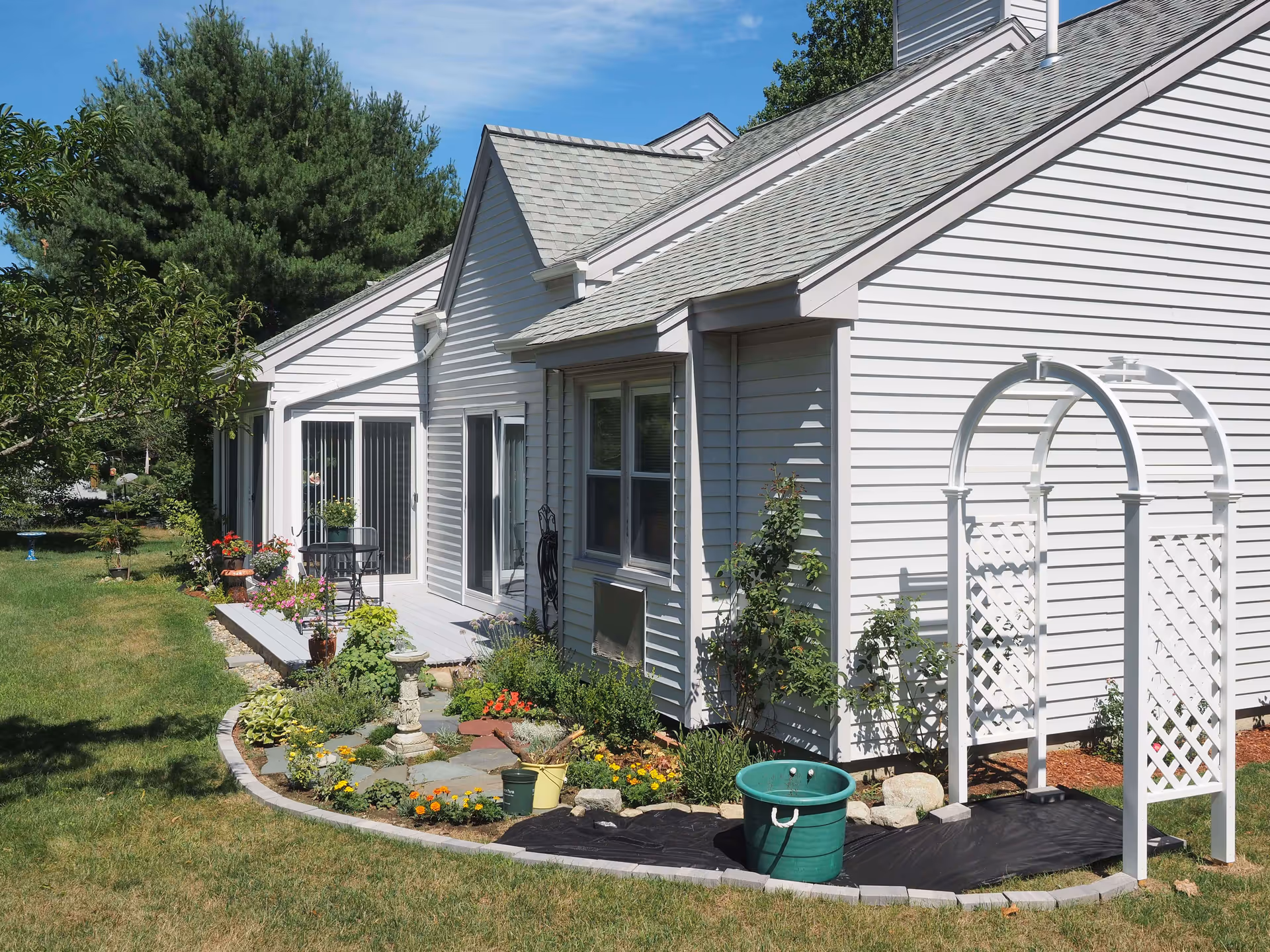 Exterior view of a white single-story building with a gray shingled roof, surrounded by a well-maintained garden with various plants and flowers. There is a small patio area with a table and chairs, a white garden arch, and a green bucket on a black tarp in the garden bed. The sky is clear and blue.