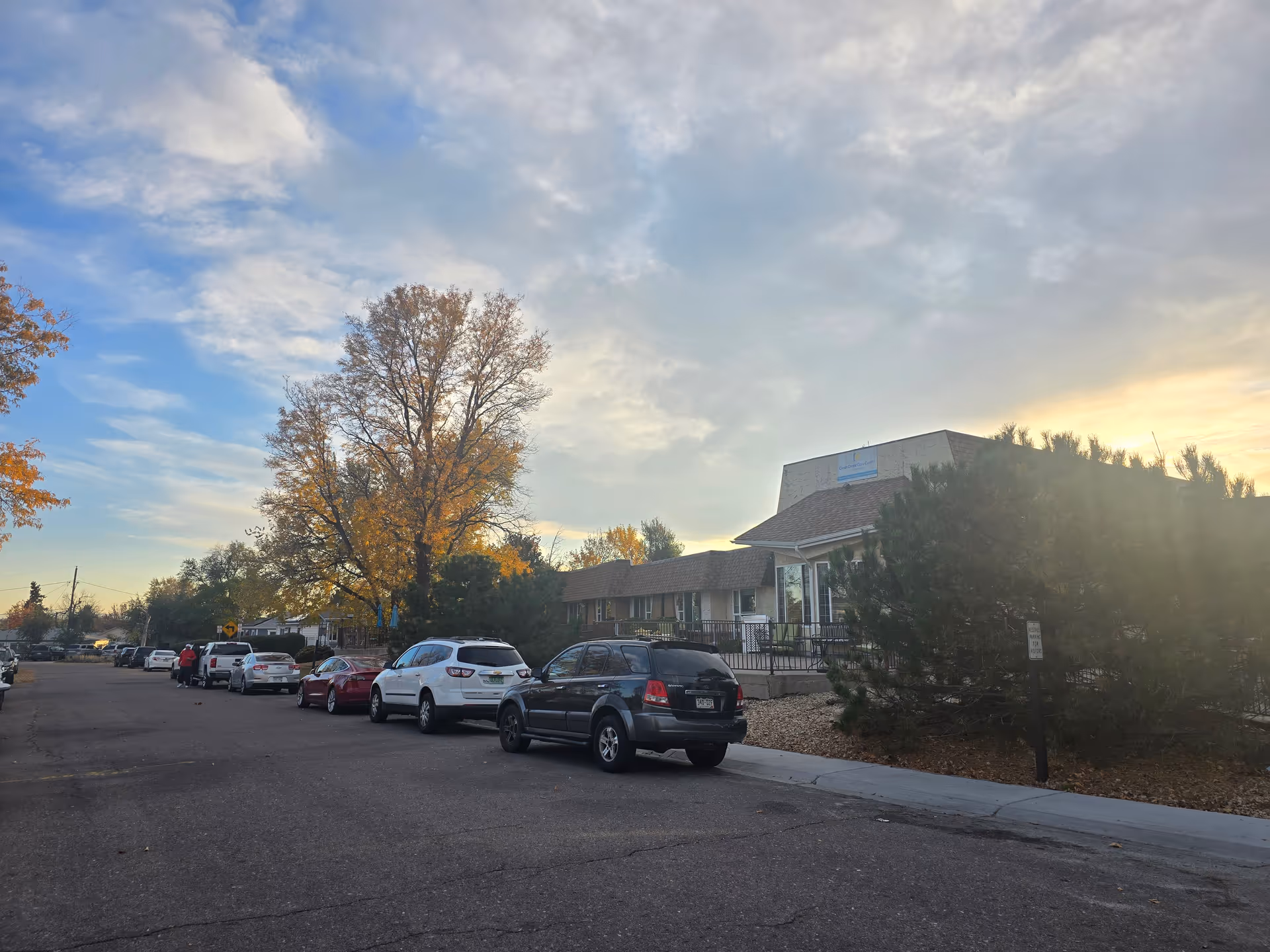Exterior view of Clear Creek Care Center building with several cars parked along the street. Trees with autumn foliage are visible, and the sky is partly cloudy with the sun setting.