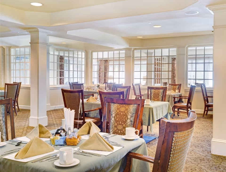 A bright and elegant dining room with multiple tables covered in light green tablecloths, each set with folded beige napkins, white cups and saucers, and silverware. The room features wooden chairs with patterned upholstery, large windows with blinds, and white pillars separating sections of the space.