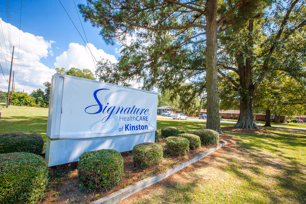 Outdoor view of the Signature HealthCARE of Kinston sign surrounded by trimmed bushes and large trees, with a clear blue sky and some clouds in the background.