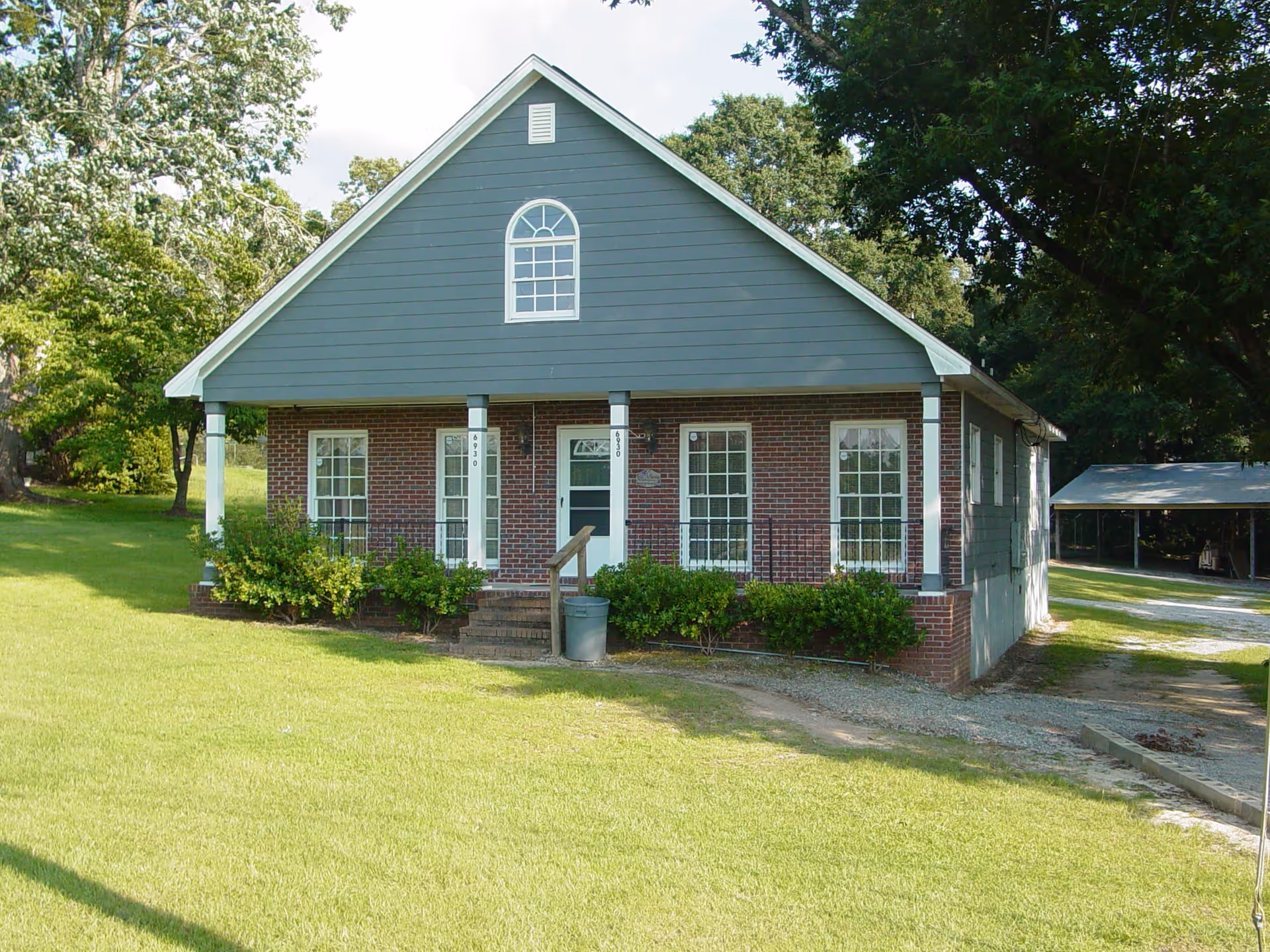 Front exterior view of a single-story brick and siding building with a gable roof, surrounded by green grass and trees. The building has four large windows and a central door with steps leading up to it. There is a gravel driveway to the right side and a covered parking area in the background.