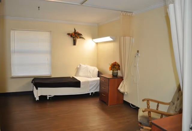 A simple nursing facility room with a single bed covered with white and black bedding, a wooden nightstand with a flower pot, a wooden rocking chair, a window with closed blinds, and a wall-mounted light above the bed. The room has beige walls and wooden flooring.