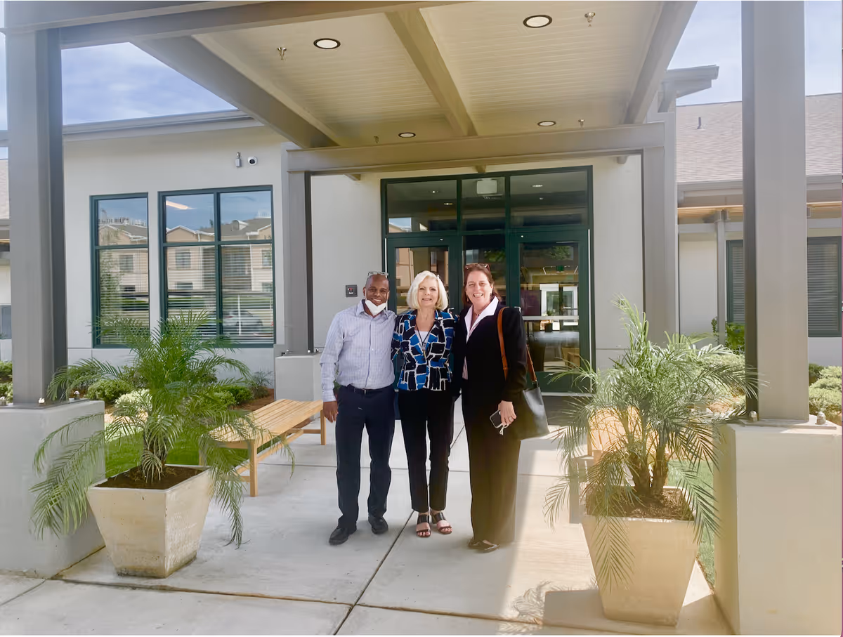 Three people standing together outside the entrance of a building with large windows and a covered walkway. There are two large potted plants on either side of the walkway and benches along the wall.