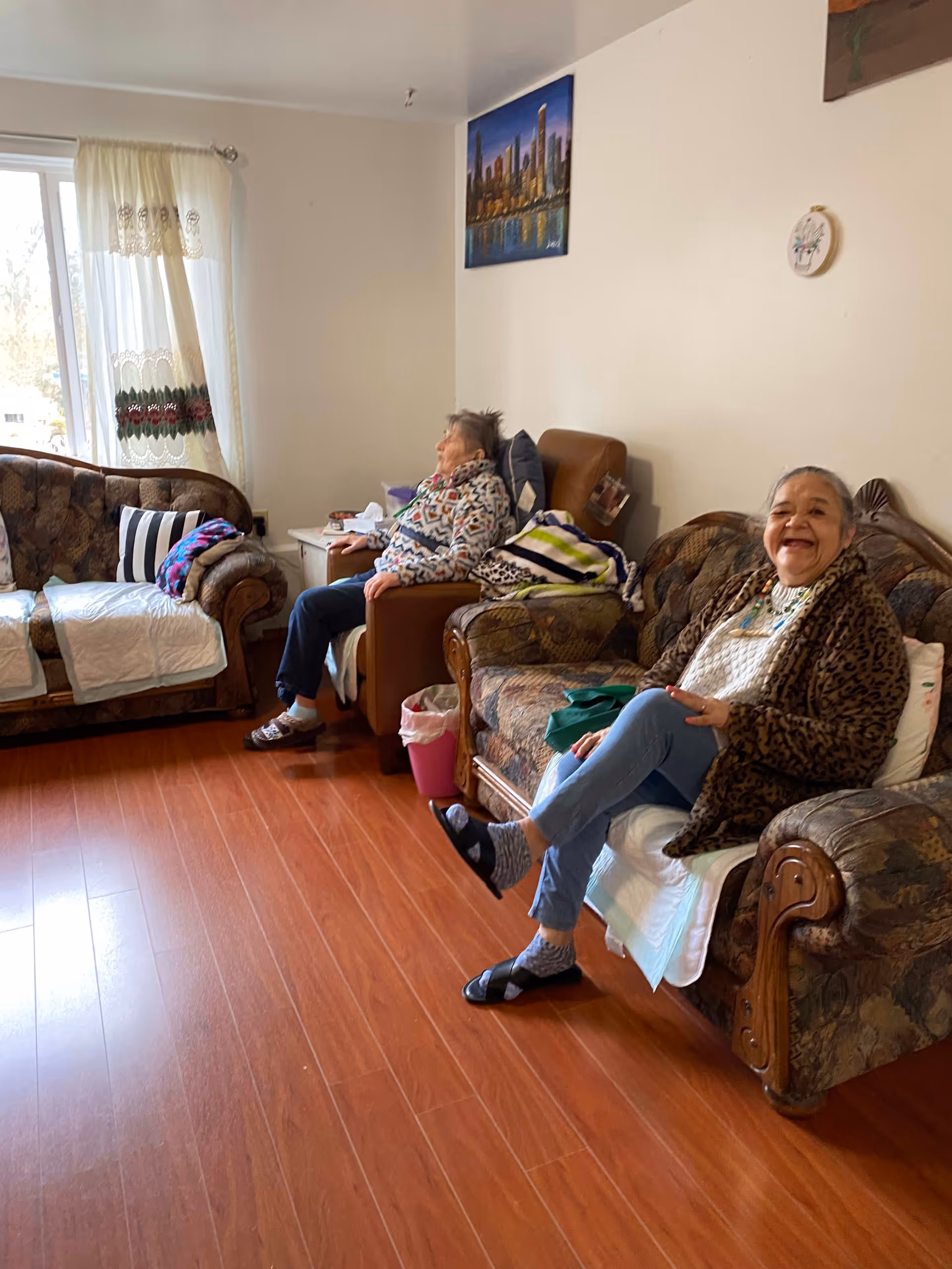 Two elderly women sitting on separate couches in a living room with wooden flooring. One woman is smiling and looking at the camera, while the other is sitting quietly looking away. The room has a window with sheer curtains, a small side table with items on it, and paintings on the wall.