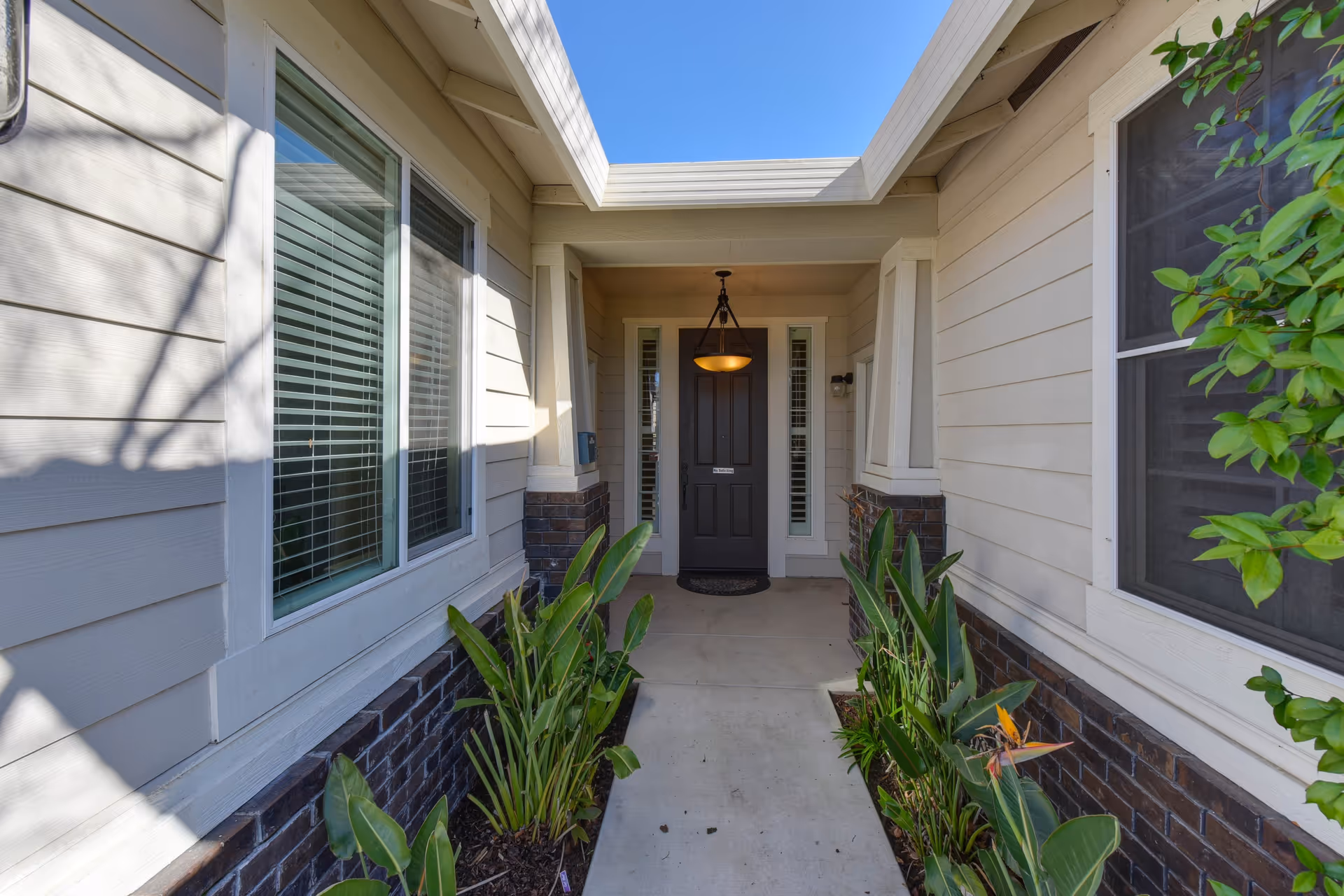 Entryway of a senior living facility with a covered porch leading to a dark front door. The walkway is flanked by green plants and the exterior walls are light-colored with brick accents. A hanging light fixture is above the door, and windows with blinds are on either side.