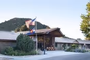 Exterior view of a single-story building with a sloped roof, surrounded by greenery and a mountain in the background. Two flagpoles display the American flag and another flag near the entrance.
