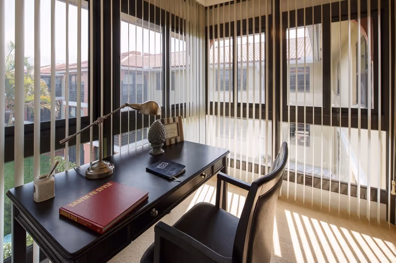 A corner office space with large windows covered by vertical blinds, a black desk with a desk lamp, decorative items, and books, and a black leather chair. Outside the windows, parts of a building with red-tiled roofs and palm trees are visible.