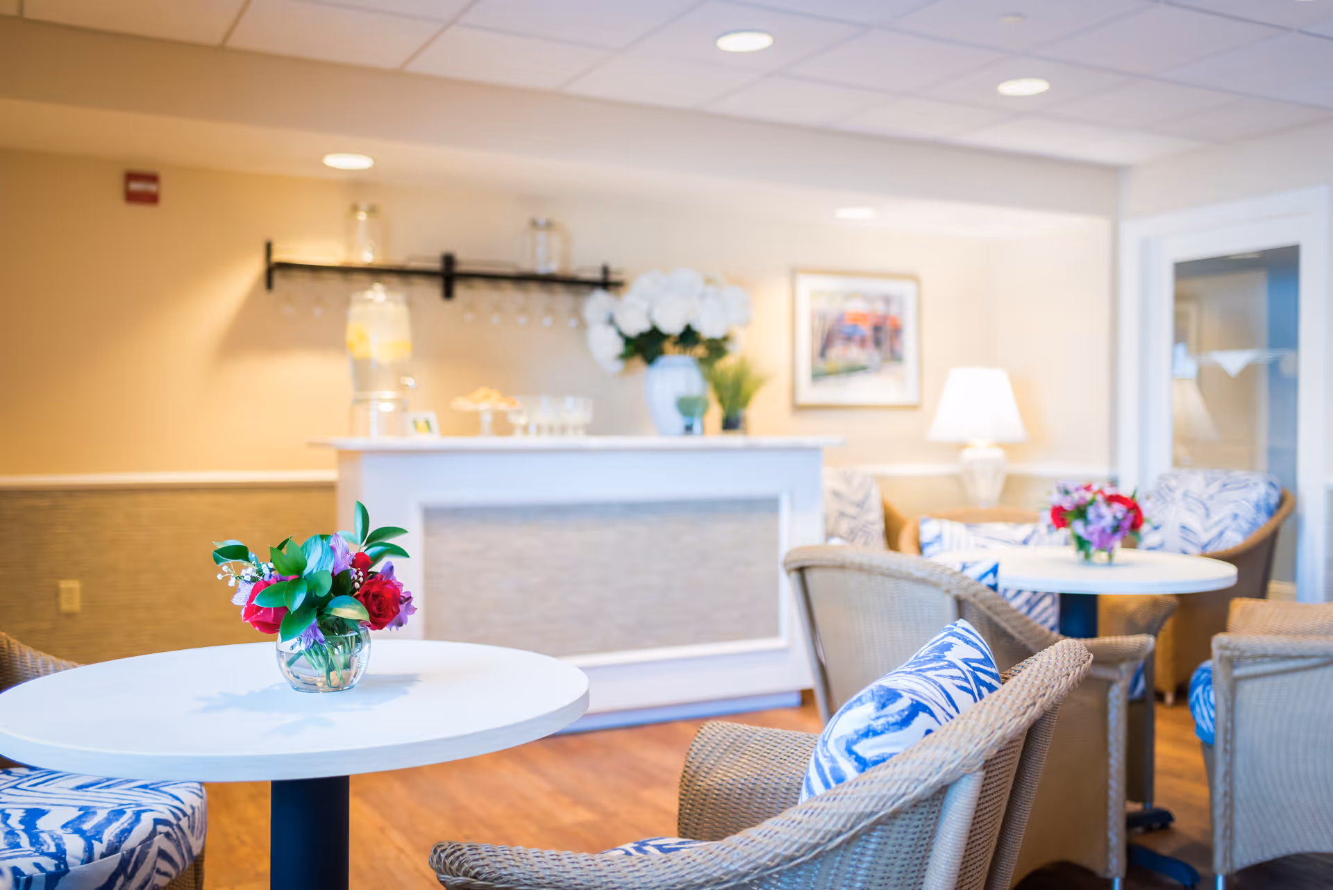 Bright common dining area with round tables, wicker chairs with patterned cushions, floral centerpieces, and a service counter in the background.