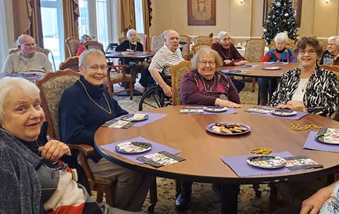 Several elderly residents seated around round tables in a decorated communal dining/activity room with a Christmas tree in the background.