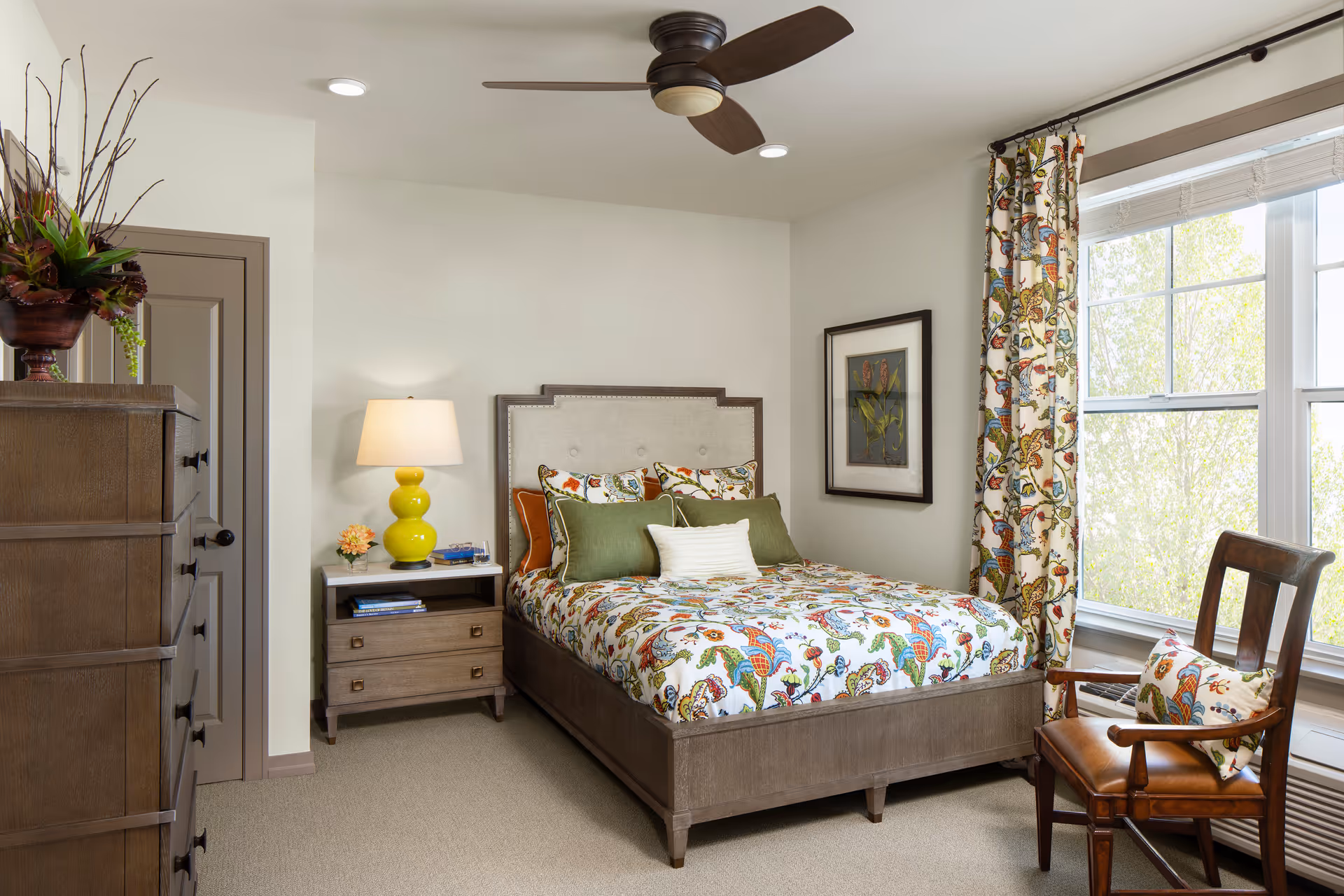 Bright bedroom with patterned bedding, a nightstand and lamp, dresser, ceiling fan, and a window with floral curtains.