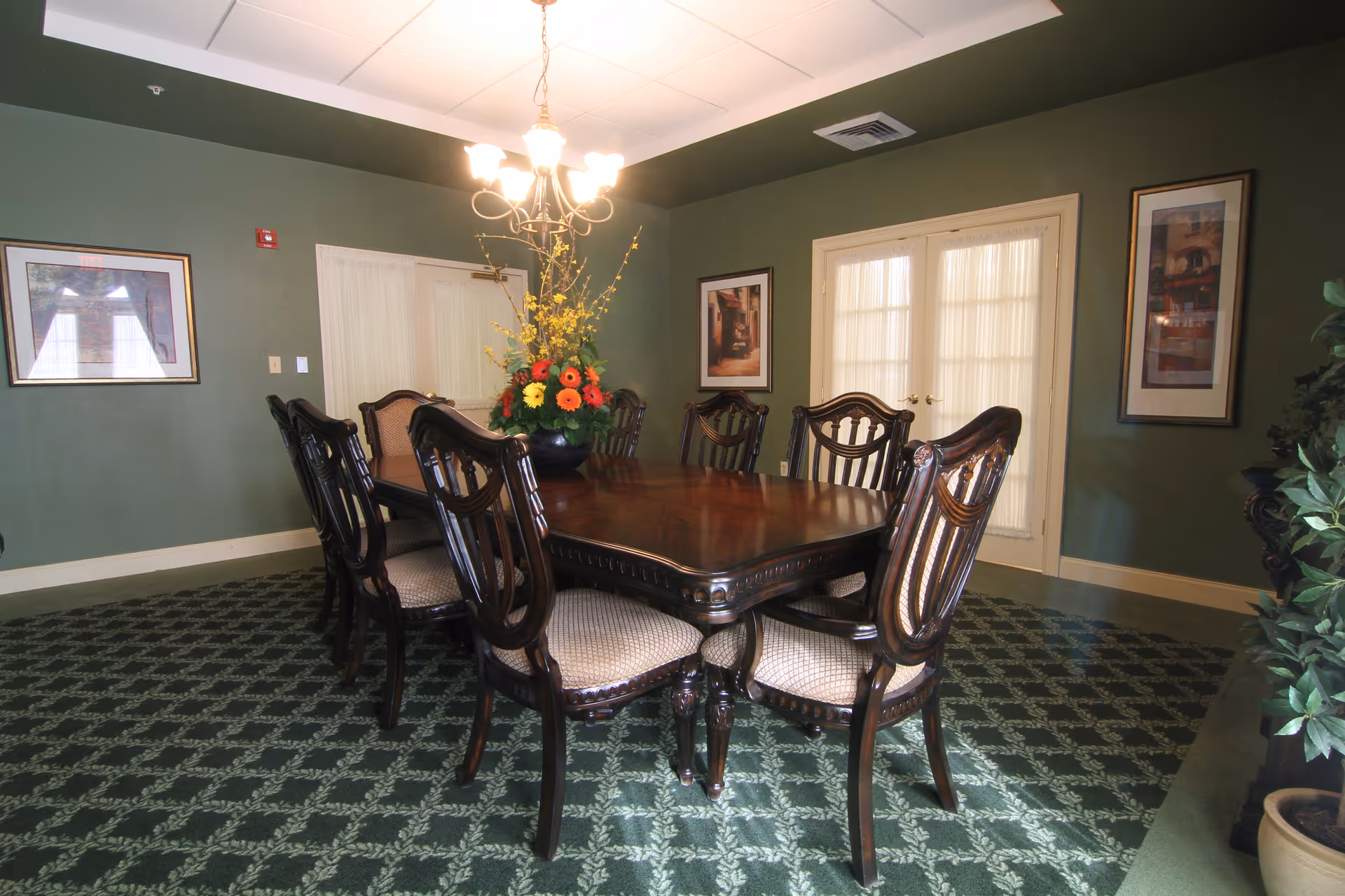 A formal dining room with a dark wooden rectangular table surrounded by eight matching chairs with cushioned seats. A floral centerpiece with yellow and orange flowers is on the table. The room has green walls, patterned green carpet, framed artwork on the walls, and double doors with sheer curtains. A chandelier with multiple lights hangs above the table.