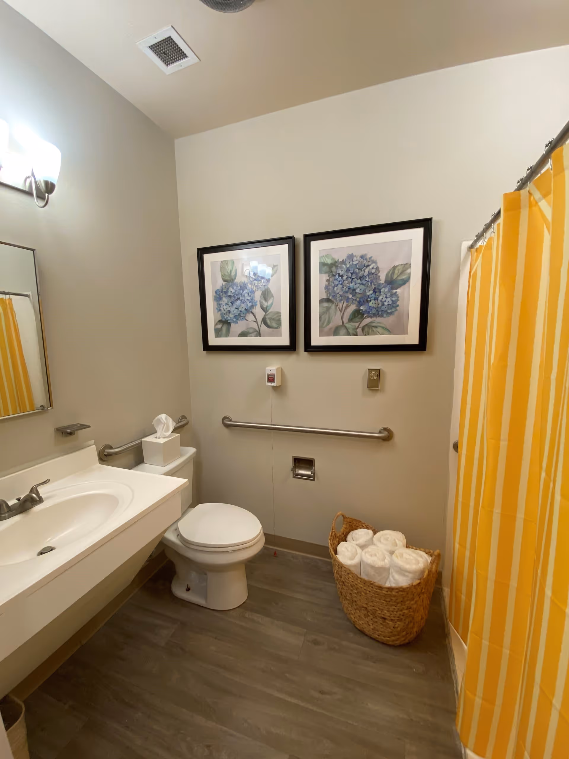A clean bathroom with a white sink and countertop, a toilet, and a yellow and white striped shower curtain. Two framed pictures of blue flowers hang on the wall above a safety grab bar. A wicker basket filled with rolled white towels is placed on the floor next to the shower.