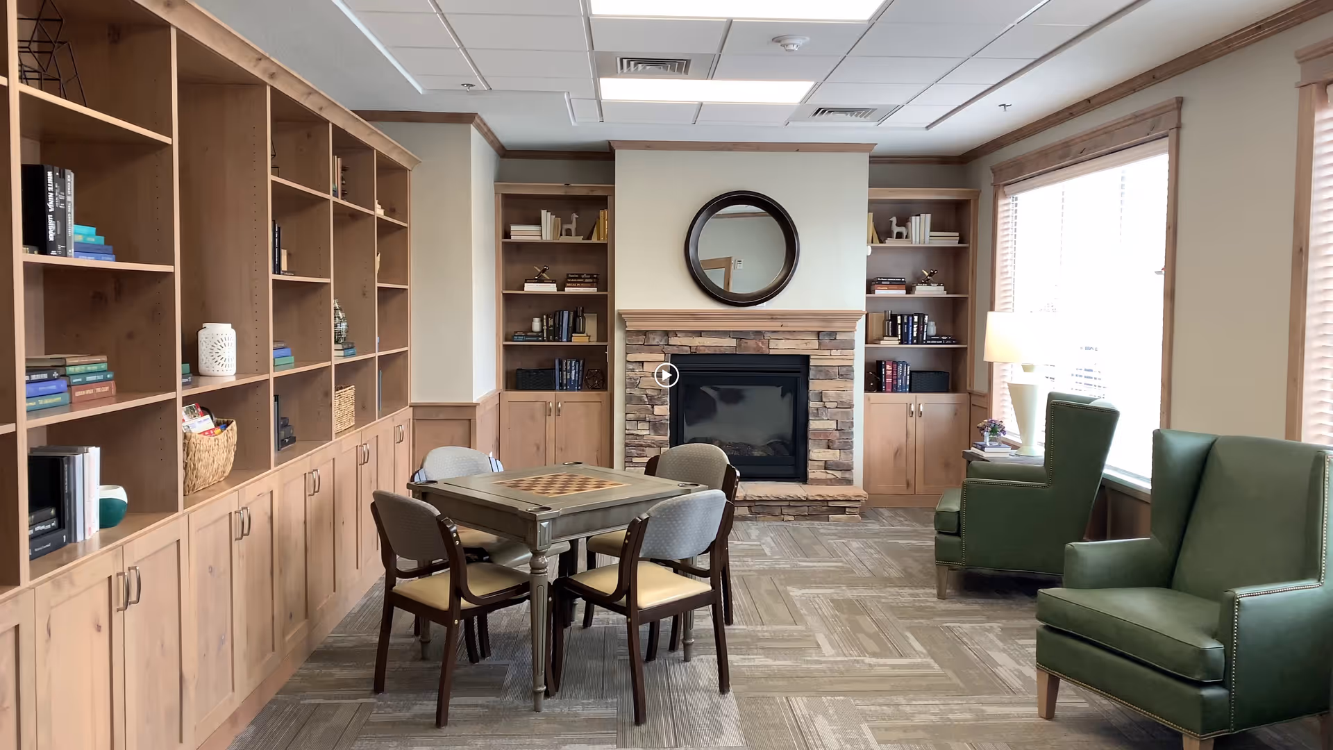 A cozy common room with a stone fireplace centered on the back wall, flanked by built-in wooden bookshelves filled with books and decorative items. In front of the fireplace is a square table with a checkerboard pattern on top, surrounded by four chairs. To the right, two green armchairs are positioned near large windows with blinds, next to a small table with a lamp and a plant. The room has a neutral color palette with wood trim and carpeted flooring.