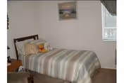 A bedroom with a bed covered in a striped bedspread, several pillows including a teddy bear, a nightstand with a lamp, a framed picture on the wall, and a window with blinds partially covered by a curtain.