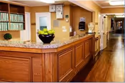 Reception area with a wooden front desk topped with a granite countertop holding a bowl of green apples. The area has warm beige walls, wooden flooring, and a hallway extending to the right with doors and wall-mounted fixtures.