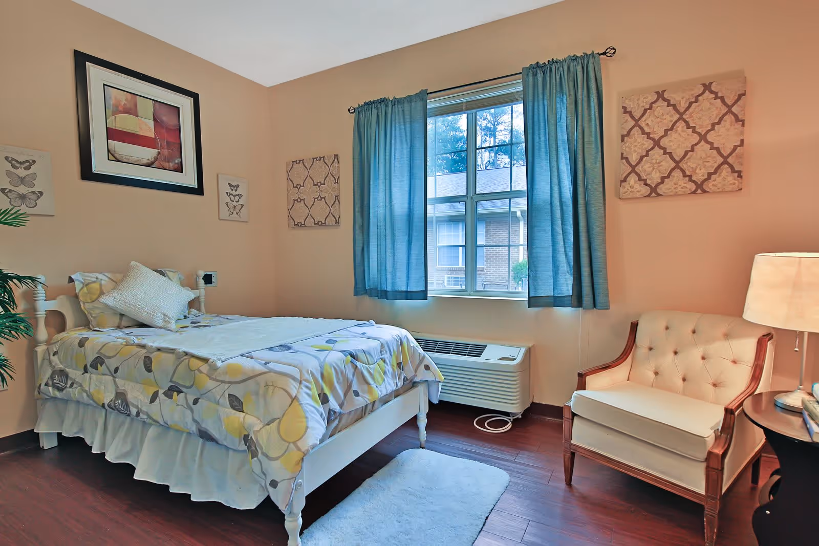 A cozy bedroom with a single bed covered in a patterned comforter and a white pillow. The room has beige walls adorned with framed artwork and butterfly prints. A window with blue curtains lets in natural light, and below it is a white air conditioning unit. To the right of the bed is a cushioned armchair with wooden arms and legs, next to a small round table with a lamp and books. The floor is wooden with a small white rug beside the bed.