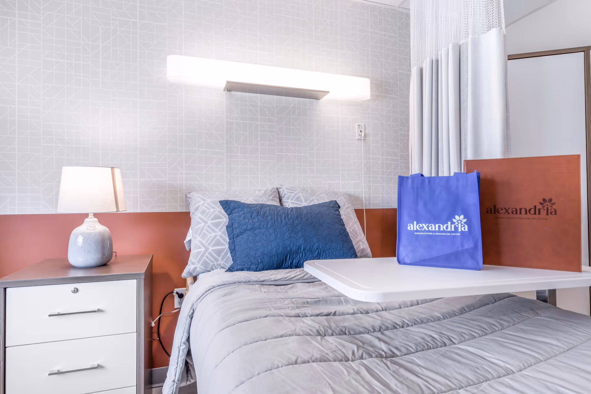 A neatly made bed with gray bedding and a blue pillow in a healthcare facility room. Next to the bed is a nightstand with a lamp. A white overbed table holds a blue tote bag and a brown folder, both branded with the name 'alexandria'. The wall behind the bed has a geometric pattern and a bright horizontal light fixture above the headboard.